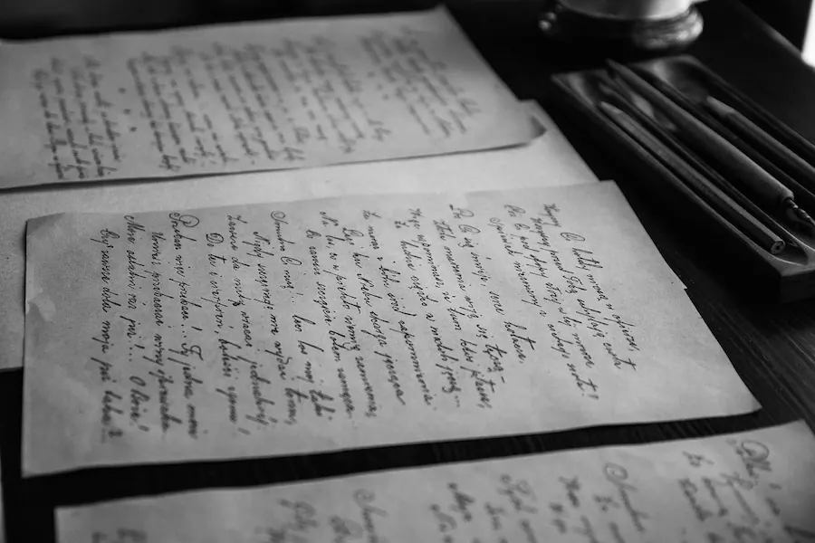 Sheets of handwritten letters laid across a dark wooden table beside a tray of pens